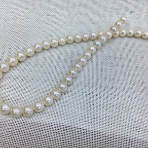 May include: A strand of white pearls, varying in size, arranged in a curved shape against a textured, off-white background. The pearls have a luminous sheen, suggesting a classic jewelry piece.