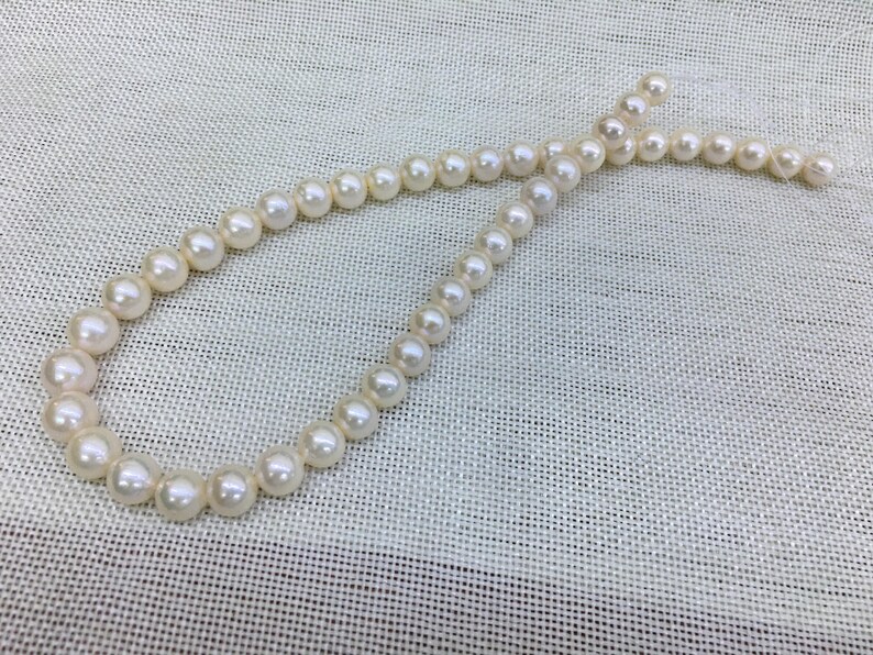 May include: A strand of white pearls, varying in size, arranged in a curved shape against a textured, off-white background. The pearls have a luminous sheen, suggesting a classic jewelry piece.