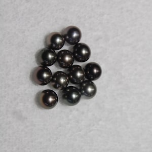 May include: A group of dark gray pearls, some with a slight iridescent sheen, are scattered on a white surface.