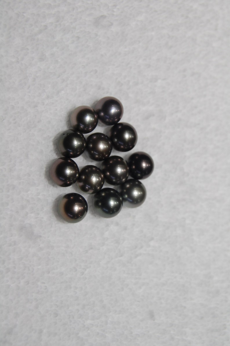 May include: A group of dark gray pearls, some with a slight iridescent sheen, are scattered on a white surface.