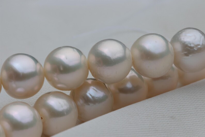 May include: Close-up of a strand of lustrous, creamy white pearls. The pearls are round and have a smooth, reflective surface. The background is a soft, off-white fabric, highlighting the pearls' natural beauty.