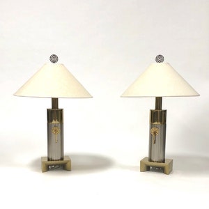 May include: A pair of silver and gold table lamps with white drum shades. Each lamp features a circular design on the base.