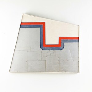 May include: Abstract art piece with a white background and a geometric design of red and blue lines. The piece is made of metal and has a textured surface.