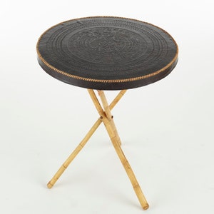 May include: A round, black leather-topped table with a decorative design on the top. The table has a tripod base made of natural bamboo.