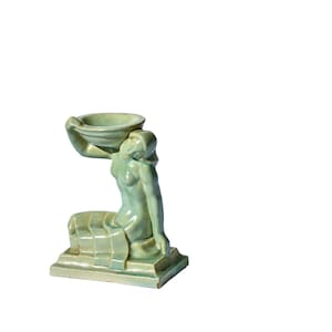 May include: A green ceramic figurine of a woman holding a bowl on her head. The figurine is sitting on a base with a rectangular shape.