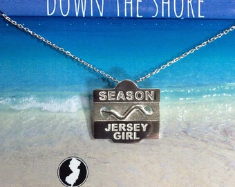 May include: Sterling silver necklace with a pendant that says "Season Jersey Girl" and a wave design. The pendant is on a silver chain against a blue and white beach background. The text "Down the Shore" is at the top of the image. The text "Jersey State Line" is at the bottom left of the image.
