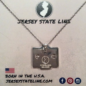 May include: Silver necklace with a pendant shaped like the state of New Jersey. The pendant is engraved with "Sterling Silver" and a smaller outline of the state. The necklace is on a white background with the text "Jersey State Line" and a website address.