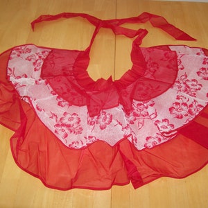 May include: A red apron with a floral pattern. The apron has a ruffled design and a tie-around neck. The apron is made of a sheer fabric and is likely used for cooking or crafting. The apron is red with white floral accents.