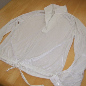 May include: A long-sleeved white shirt with a shimmering finish, featuring a pointed collar, a deep V-neckline, and a drawstring hem with ties. The sleeves have buttoned cuffs, and the fabric has a subtle, reflective, metallic-like sheen.