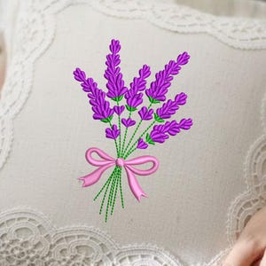 Puede incluir: Cojín cuadrado color crema con bordado de ramo de lavanda. El ramo presenta flores de lavanda moradas, tallos verdes y un lazo rosa. El cojín tiene un delicado ribete de encaje en los bordes.
