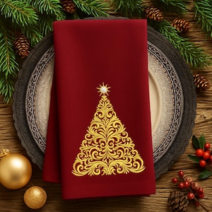 May include: A folded, deep red cloth napkin with a gold embroidered Christmas tree design. The tree features intricate swirling patterns and is topped with a star. The napkin is placed on a decorative plate, with pine needles and ornaments in the background.