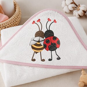 Puede incluir: Toalla de bebé blanca con capucha y ribete rosa. La toalla presenta un diseño bordado de una abeja y una mariquita de dibujos animados abrazándose. La abeja es amarilla y marrón, y la mariquita es roja con lunares negros.