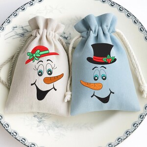 May include: Two drawstring gift bags with embroidered snowman faces. One bag is beige with a red and green hat, the other is light blue with a black top hat and holly. Both have orange carrot noses and smiling mouths.