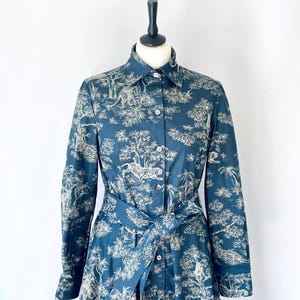 May include: A long-sleeved, button-down dress in a dark teal color, featuring a white toile print of tigers, trees, and foliage. The dress has a matching tie belt at the waist and is displayed on a mannequin against a white background.