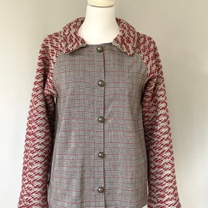 Puede incluir: Chaqueta abotonada con frente a cuadros y mangas texturizadas. La parte delantera de la chaqueta presenta un patrón de cuadros grises, rojos y negros. Las mangas y el cuello son de punto texturizado rojo y gris. La chaqueta se exhibe en un maniquí blanco.