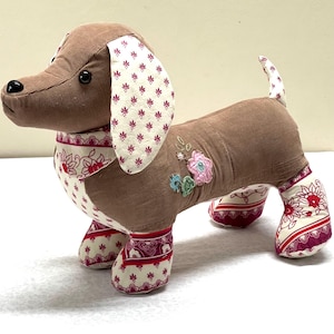May include: A stuffed dachshund dog with a brown corduroy body, cream-colored ears and paws with a burgundy floral pattern. The dog has embroidered flowers on its side and a burgundy and cream patterned collar.