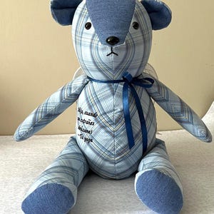 May include: A stuffed teddy bear crafted from light blue plaid fabric with solid blue denim accents on the ears, snout, and feet. A dark blue ribbon is tied around its neck. The chest features embroidered text: Mija, cuando me extrañes abrázame! Tu papa.