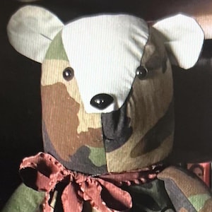 May include: A stuffed bear toy featuring a camouflage pattern in shades of green, brown, and tan. The bear has a white fabric patch on its face, two small black bead eyes, a black nose, and a dark red ribbon tied around its neck.