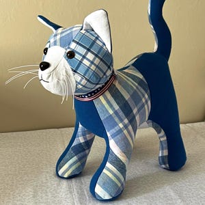 May include: A stuffed cat figurine crafted from blue and white plaid fabric and solid blue fabric. The cat features a white face, dark button eyes, a black nose, and white whiskers. It wears a collar with a red, white, and blue star pattern.