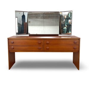 May include: A teak wood vanity table featuring a rectangular base with four drawers, each fitted with two circular wooden knobs. A large tri-fold mirror with three distinct glass panels is attached to the top surface of the wooden dresser unit.