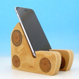 May include: A wooden phone stand with a natural wood finish. The stand has a triangular base with three circular cutouts and a raised platform for holding a phone.