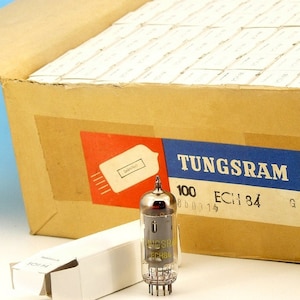 Puede incluir: Una caja de 100 tubos de vacío TUNGSRAM ECH 84. La caja es marrón con una etiqueta roja y azul. La etiqueta dice "TUNGSRAM 100 ECH 84". Se muestra un solo tubo de vacío delante de la caja.