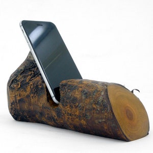 Puede incluir: Un soporte de teléfono de madera hecho de una rama de árbol con un acabado de corteza natural. Un teléfono inteligente negro descansa en el soporte.