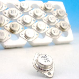Puede incluir: Primer plano de un transistor plateado con el texto "dai OC 29 9042" impreso en él. El transistor está sobre una superficie blanca.