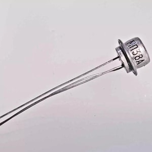 May include: A close-up of an electronic component, a transistor, with three metal leads extending from a silver-colored, circular body. The text "MT38A" is printed on the top of the component. The background is a plain white surface.