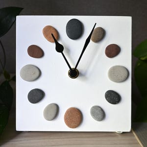 Könnte beinhalten: Eine quadratische, weiße Wanduhr mit zwölf glatten, mehrfarbigen Flusskieselsteinen, die kreisförmig als Stundenmarkierungen angeordnet sind. Die Uhr hat zwei dünne, schwarze Metallzeiger in der Mitte, die auf elf und eins zeigen.