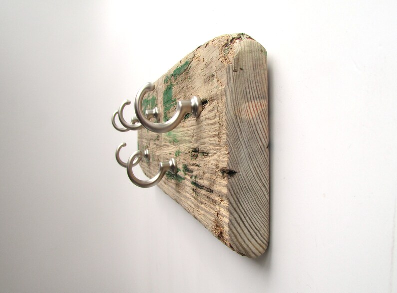 Op de afbeelding: Een rustieke houten kapstok met zes zilveren metalen haken. Het hout is verweerd met zichtbare nerven en groene verfvlekken. De haken zijn gelijkmatig verdeeld en gebogen, wat een praktische opbergoplossing biedt.