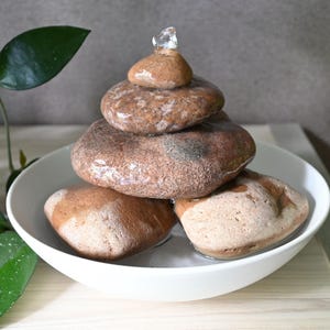 Pode incluir: Uma fonte de água de mesa com uma pilha de pedras castanhas lisas e arredondadas dispostas em forma de pirâmide dentro de uma taça de cerâmica branca, rasa e circular. Um pequeno jato de água flui do topo da pilha para a base.