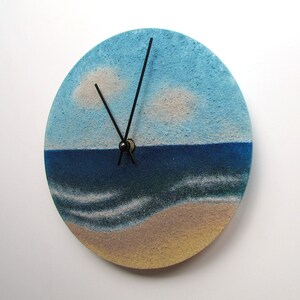 Pu&ograve; includere: Un orologio rotondo con un design di cielo blu, nuvole bianche, oceano blu e spiaggia sabbiosa. Le lancette dell'orologio sono nere.