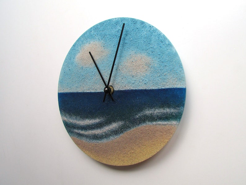 Pu&ograve; includere: Un orologio rotondo con un design di cielo blu, nuvole bianche, oceano blu e spiaggia sabbiosa. Le lancette dell'orologio sono nere.
