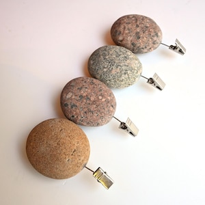 May include: Four smooth, rounded river stones arranged in a diagonal line, each featuring a unique natural speckled pattern in shades of tan, gray, and brown. Each stone is attached to a small, silver-toned metal alligator clip.