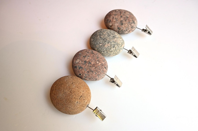 May include: Four smooth, rounded river stones arranged in a diagonal line, each featuring a unique natural speckled pattern in shades of tan, gray, and brown. Each stone is attached to a small, silver-toned metal alligator clip.