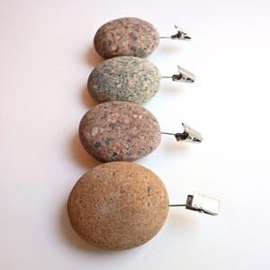 May include: Four smooth, rounded river stones arranged in a vertical column, each featuring a unique speckled texture in shades of brown, gray, and tan. Each stone is fitted with a small, silver-toned metal alligator clip attached to the side.