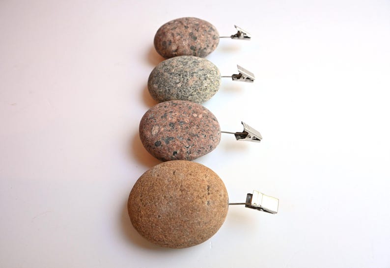 May include: Four smooth, rounded river stones arranged in a vertical column, each featuring a unique speckled texture in shades of brown, gray, and tan. Each stone is fitted with a small, silver-toned metal alligator clip attached to the side.