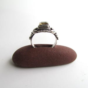 Peut inclure: Une bague en argent avec une pierre pr&eacute;cieuse jaune sertie dans une b&eacute;li&egrave;re d&eacute;corative. La bague est pr&eacute;sent&eacute;e sur un porte-bague en pierre brune.