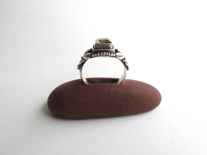 Peut inclure: Une bague en argent avec une pierre pr&eacute;cieuse jaune sertie dans une b&eacute;li&egrave;re d&eacute;corative. La bague est pr&eacute;sent&eacute;e sur un porte-bague en pierre brune.