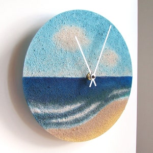 Pu&ograve; includere: Un orologio rotondo con un design a paesaggio marino blu e bianco. Il quadrante dell'orologio &egrave; realizzato in un materiale testurizzato che ricorda la sabbia. Le lancette dell'orologio sono bianche e il quadrante &egrave; circondato da un bordo marrone.