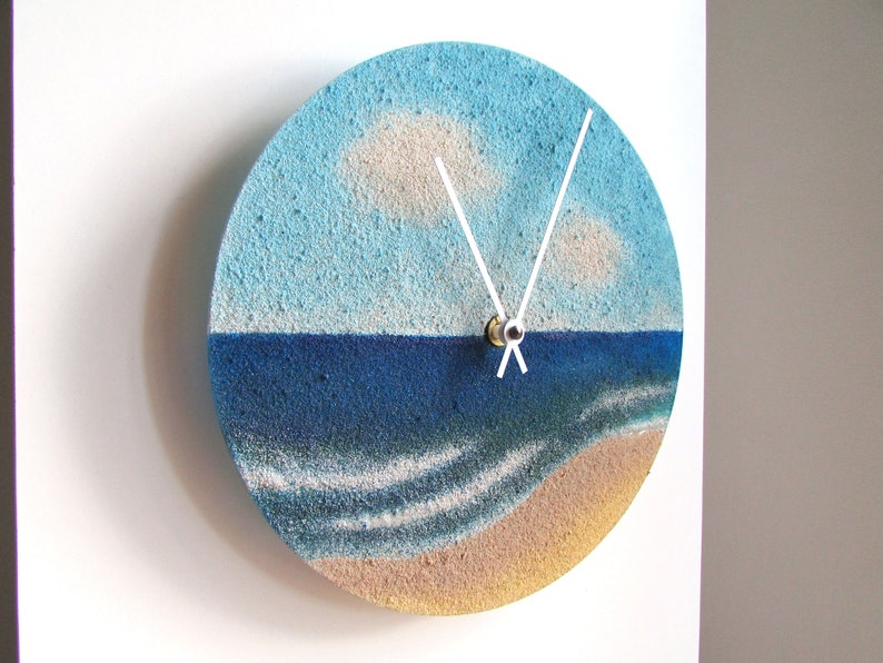 Pu&ograve; includere: Un orologio rotondo con un design a paesaggio marino blu e bianco. Il quadrante dell'orologio &egrave; realizzato in un materiale testurizzato che ricorda la sabbia. Le lancette dell'orologio sono bianche e il quadrante &egrave; circondato da un bordo marrone.