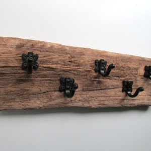 May include: A rustic wooden wall-mounted coat rack with five black metal hooks. The wood has a weathered, distressed look and the hooks have a decorative, ornate design.