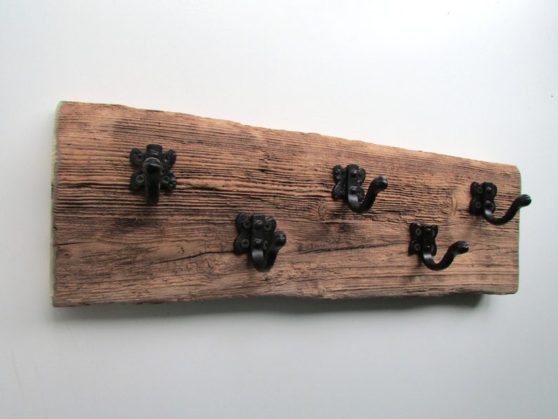 May include: A rustic wooden wall-mounted coat rack with five black metal hooks. The wood has a weathered, distressed look and the hooks have a decorative, ornate design.