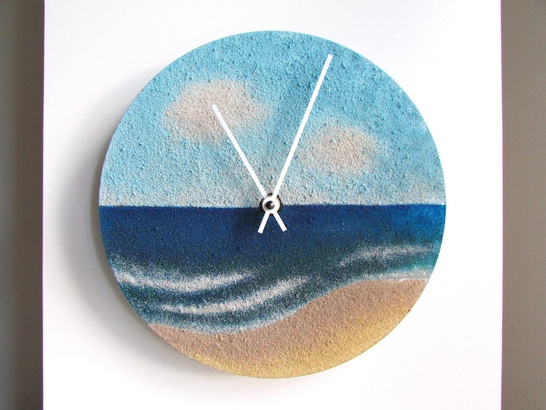 Pu&ograve; includere: Un orologio rotondo con un design a paesaggio marino blu e bianco. Il quadrante dell'orologio &egrave; dipinto per assomigliare a una scena di spiaggia con acqua blu, onde bianche e una spiaggia sabbiosa. Le lancette dell'orologio sono bianche.