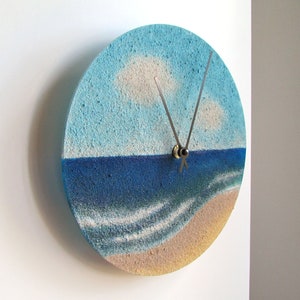 Pu&ograve; includere: Un orologio rotondo con un design a paesaggio marino blu e bianco. Il quadrante dell'orologio &egrave; realizzato in un materiale testurizzato che ricorda la sabbia. Le lancette dell'orologio sono argentate.