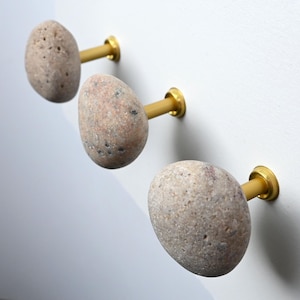 Könnte beinhalten: Drei Wandhaken mit natürlichen, abgerundeten, gesprenkelten beigen und grauen Steinknöpfen, die an zylindrischen, gebürsteten goldfarbenen Metallsockeln befestigt sind. Jeder Stein hat eine einzigartige, strukturierte Oberfläche mit kleinen Vertiefungen.