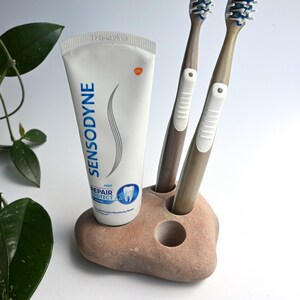 Può includere: Un tubetto bianco di dentifricio Sensodyne Repair and Protect accanto a due spazzolini marroni con impugnature in gomma bianca, inseriti in un supporto liscio color beige simile a una pietra con tre aperture circolari per gli oggetti.