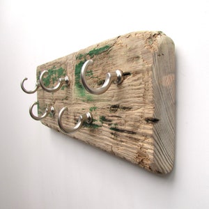 Op de afbeelding: Een rustieke houten kapstok met zes zilveren metalen haken. Het hout is verweerd met zichtbare nerven en groene verfvlekken. De haken zijn gebogen en gelijkmatig verdeeld.