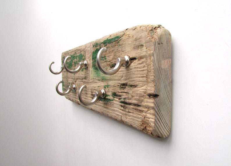 Op de afbeelding: Een rustieke houten kapstok met zes zilveren metalen haken. Het hout is verweerd met zichtbare nerven en groene verfvlekken. De haken zijn gebogen en gelijkmatig verdeeld.